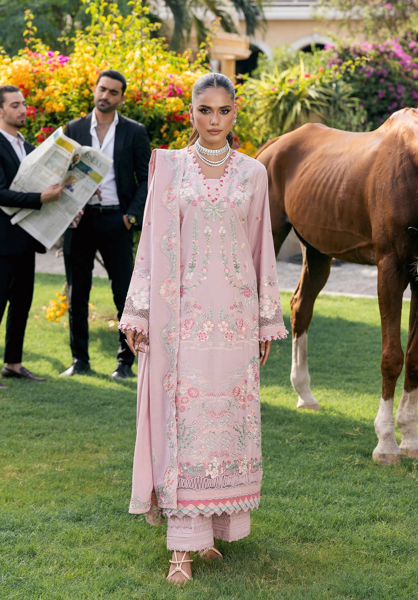 Woman in a light pink traditional outfit standing next to a horse with two men in the background.