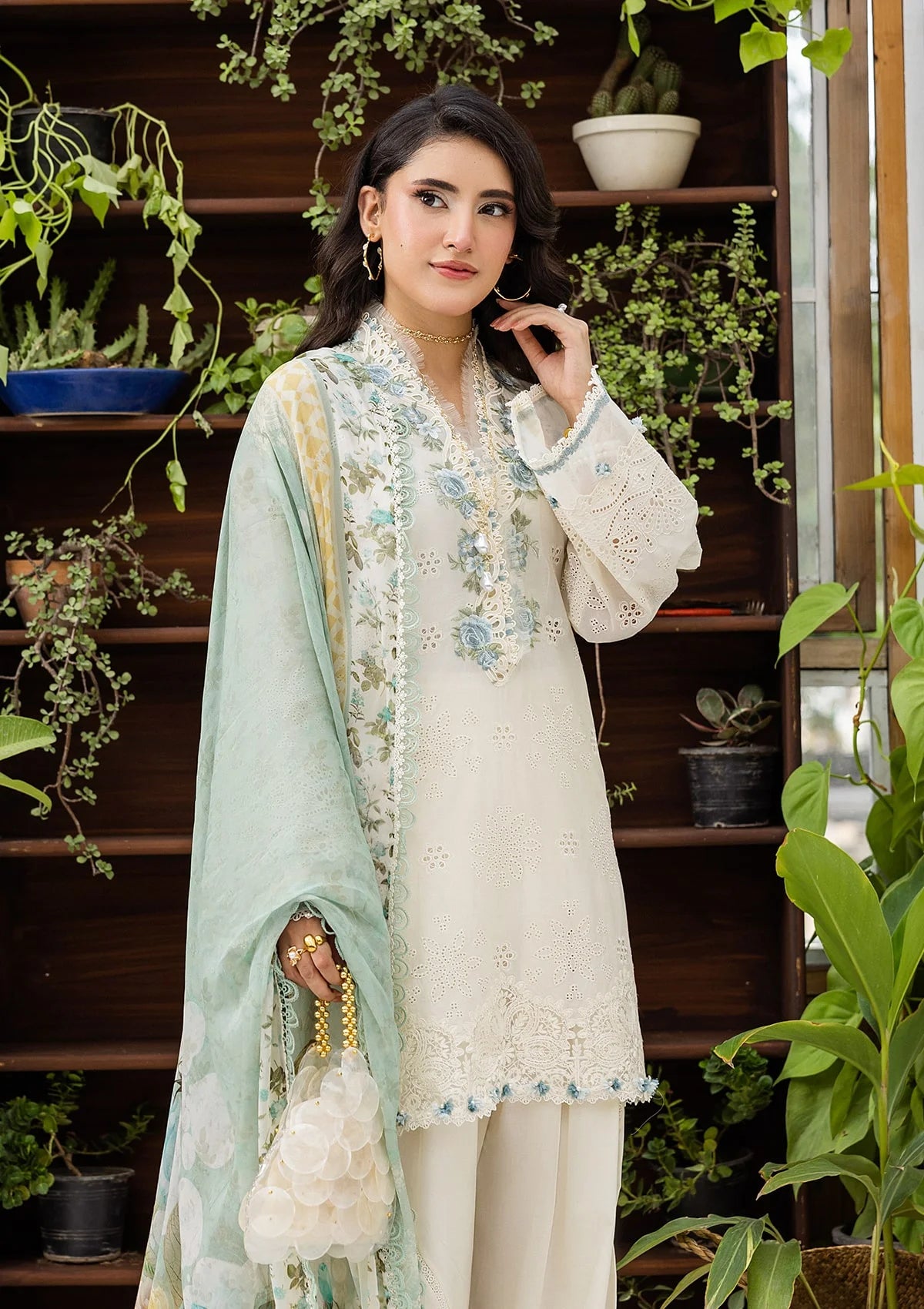 Woman in a light-colored embroidered outfit with a green dupatta, standing in a plant-filled room.