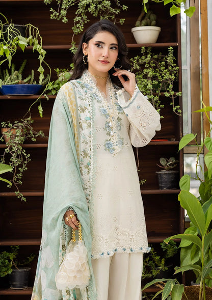Woman in a light-colored embroidered outfit with a green dupatta, standing in a plant-filled room.