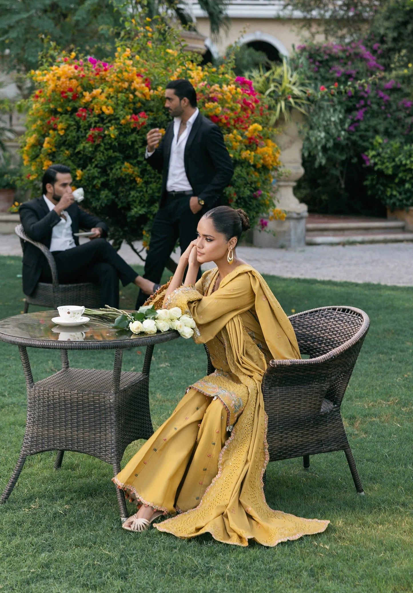 Woman in a yellow saree sitting outdoors with two men in the background.