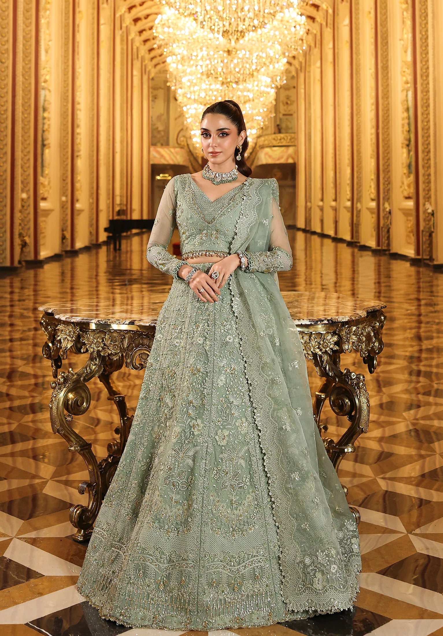 Woman in a light green embroidered traditional outfit standing in an opulent room with chandelier with with Elaf Stardom Handwork.