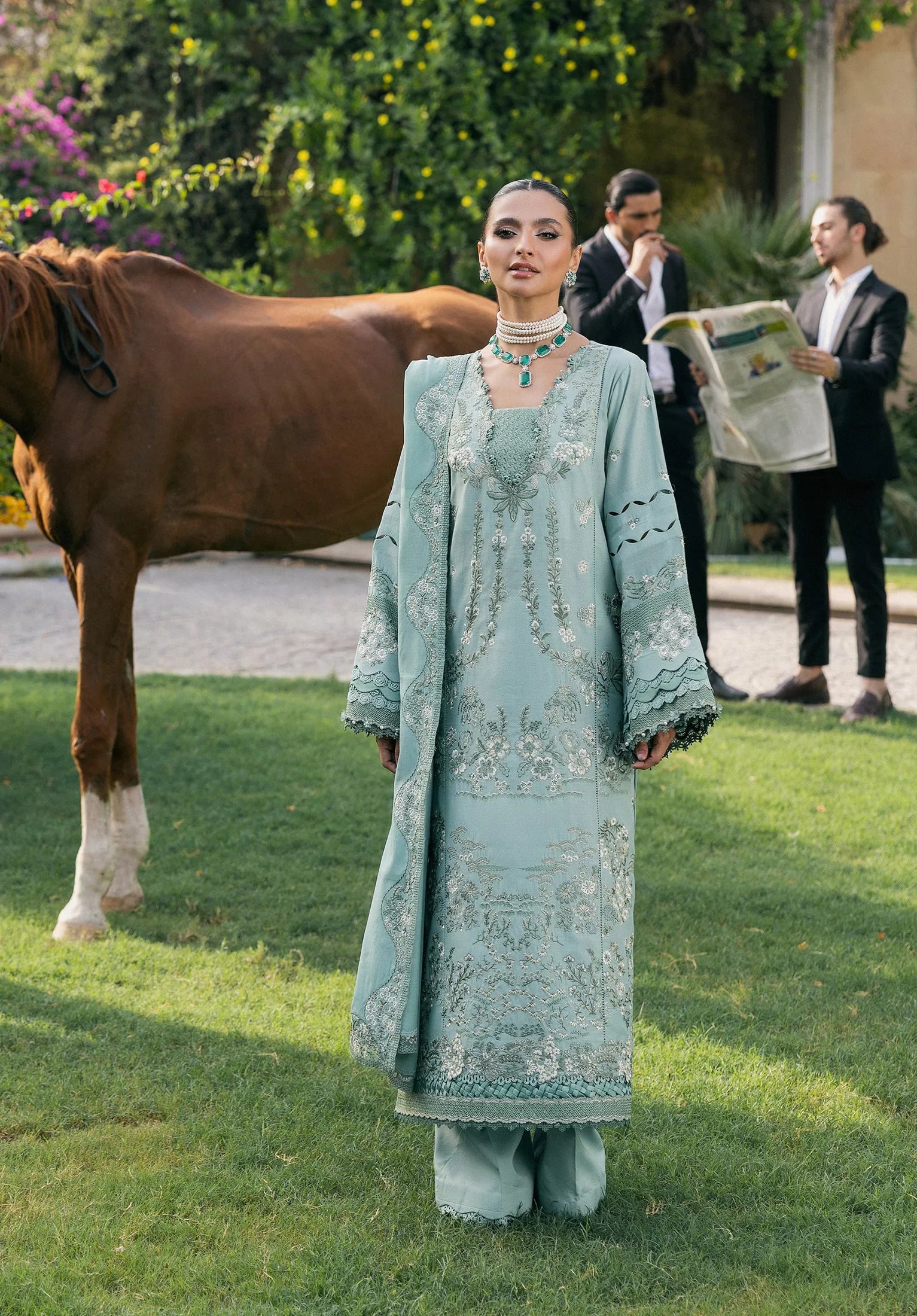 Woman in a light blue embroidered outfit standing in a grassy area with a horse and people in the background.