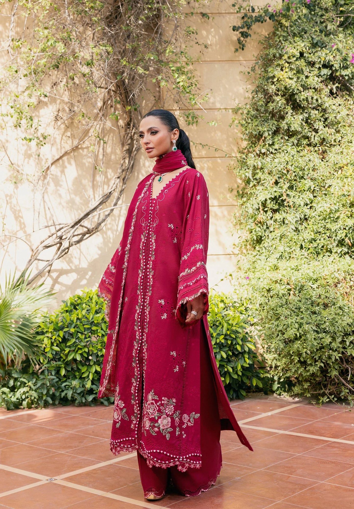 Woman in a red traditional outfit standing outdoors with greenery and stone wall in the background